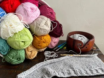 Photo of a stack of colorful yarn, knitting tools and a knitted piece in progress sitting atop a table.